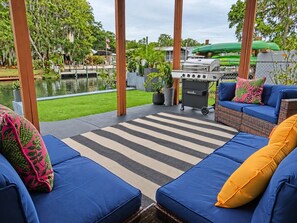 An outdoor patio with blue cushioned seating, a grill, and a view of a body of water and greenery.