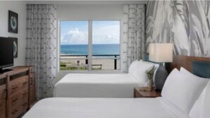 Habitación de hotel con dos camas, televisor, lámpara y vista al océano.