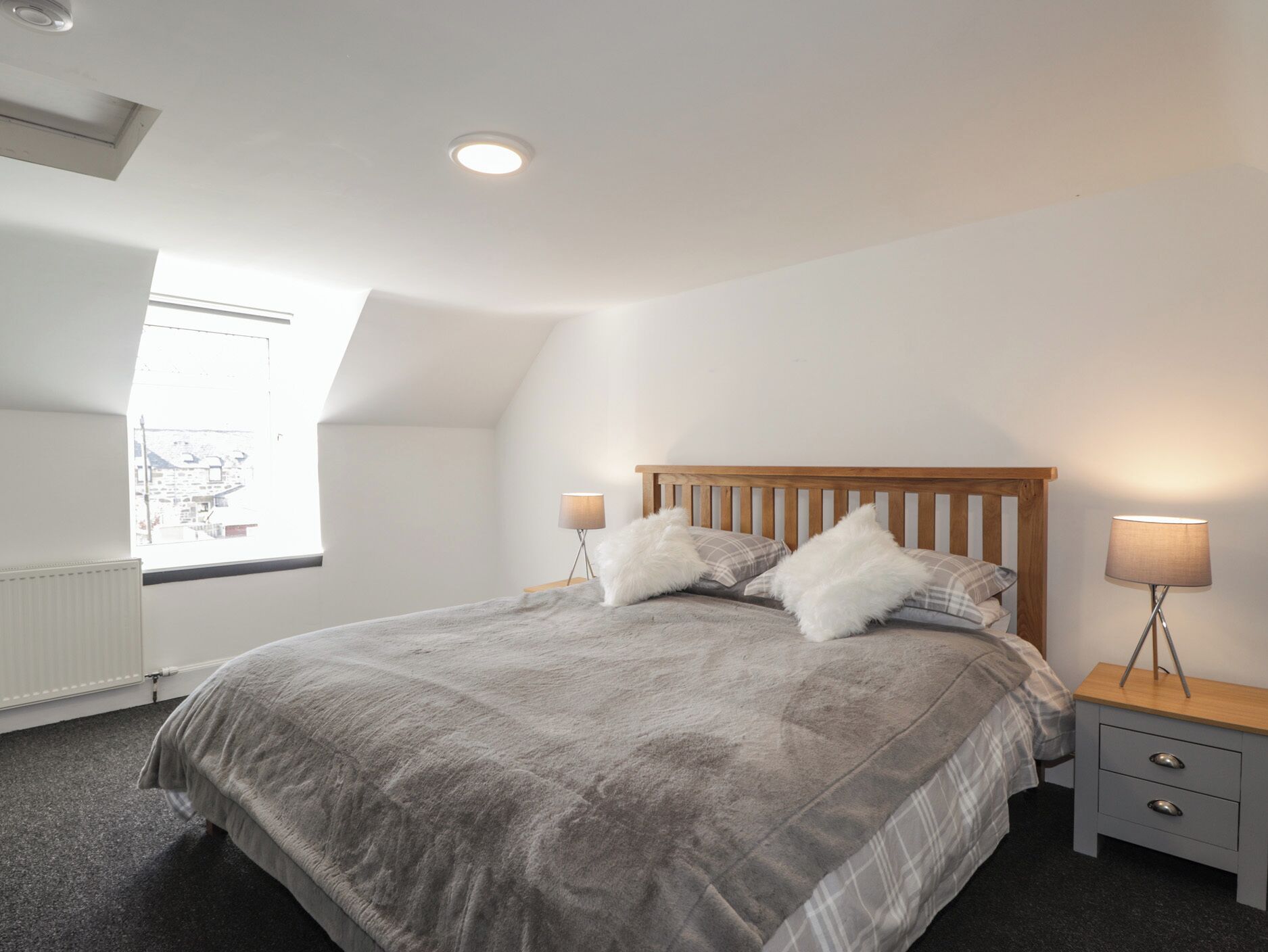 A bedroom with a sloped ceiling, a large bed with a wooden headboard, two bedside tables with lamps, and a window with a view of buildings.