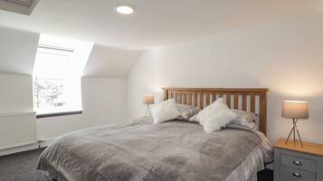 A bedroom with a sloped ceiling, a large bed with a wooden headboard, two bedside tables with lamps, and a window with a view of buildings.
