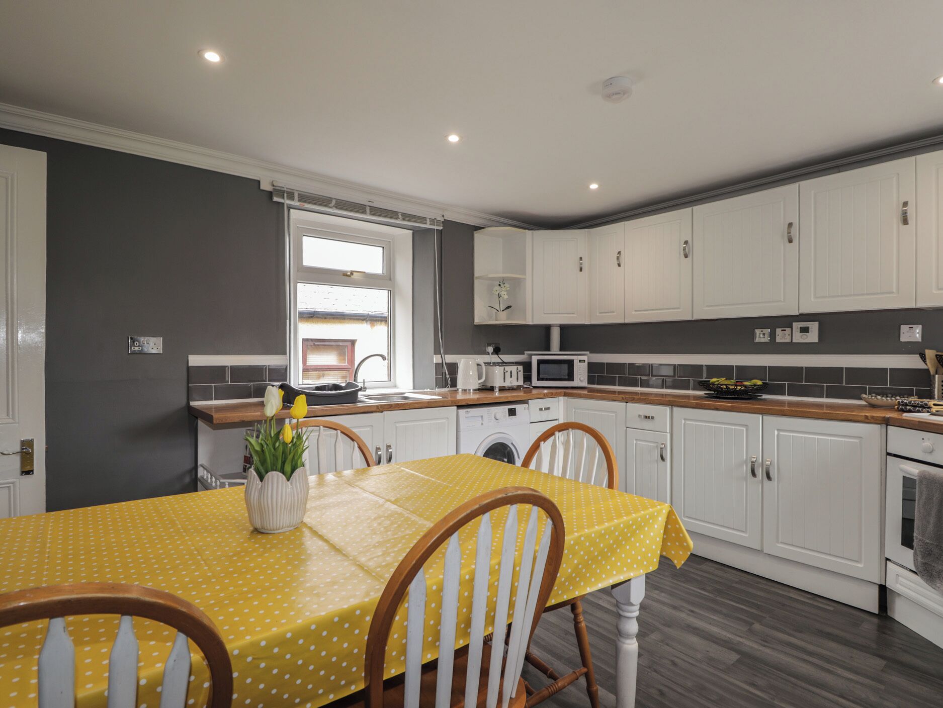 Una cocina compacta con armarios blancos, una mesa amarilla con mantel estampado de lunares y una ventana con vistas al exterior.