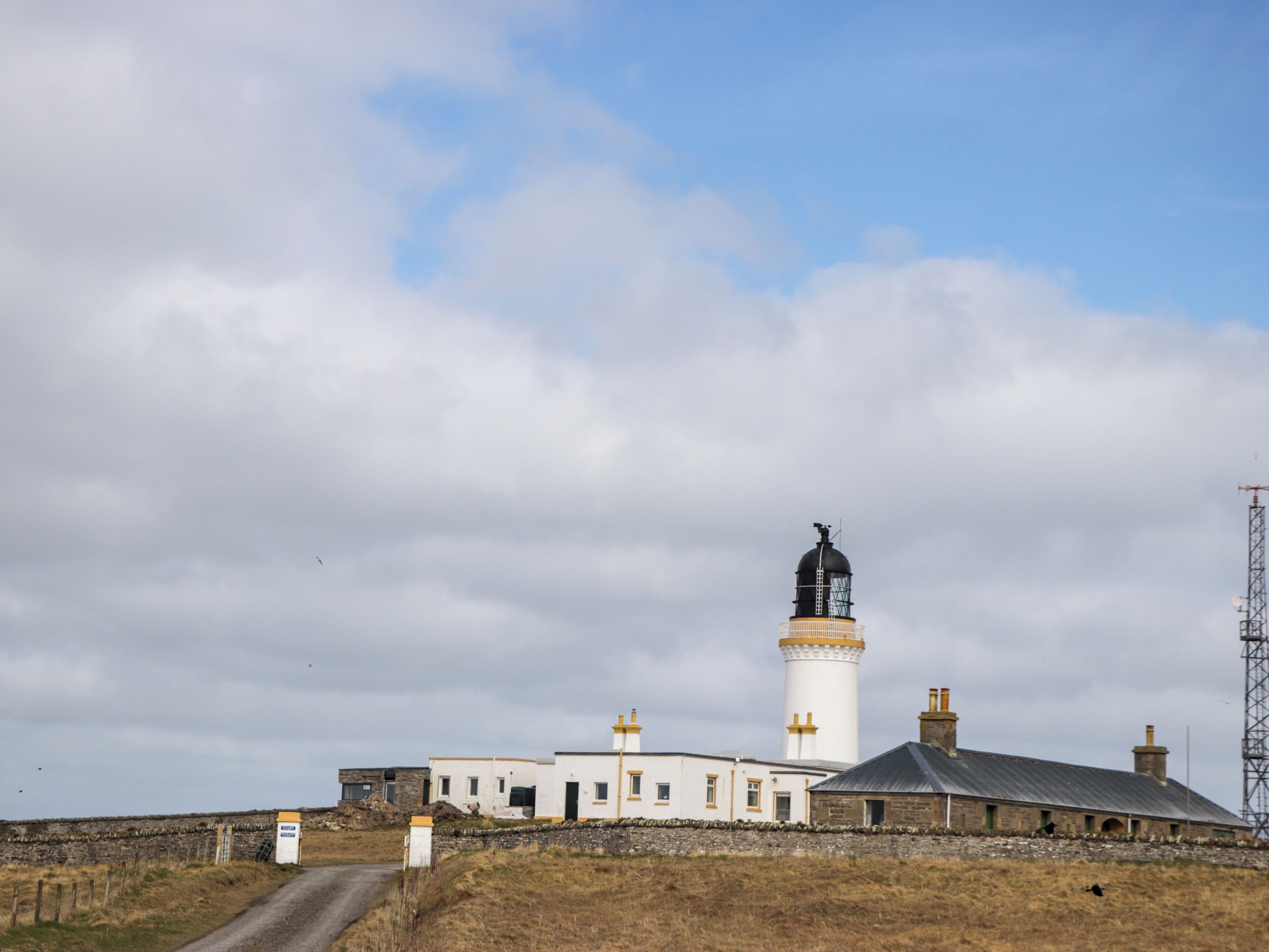 A lighthouse with a white and black tower, a small adjacent building, and a gravel path leading to the entrance.