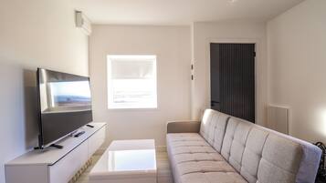 A modern living room with a flat-screen TV on a white console, a beige sofa, and a window with blinds.