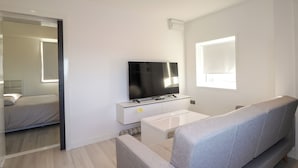 A modern living room with a flat-screen TV on a white console, a grey sofa, and a window with blinds. - The Lighthouse Keepers Cabin (Wick)
