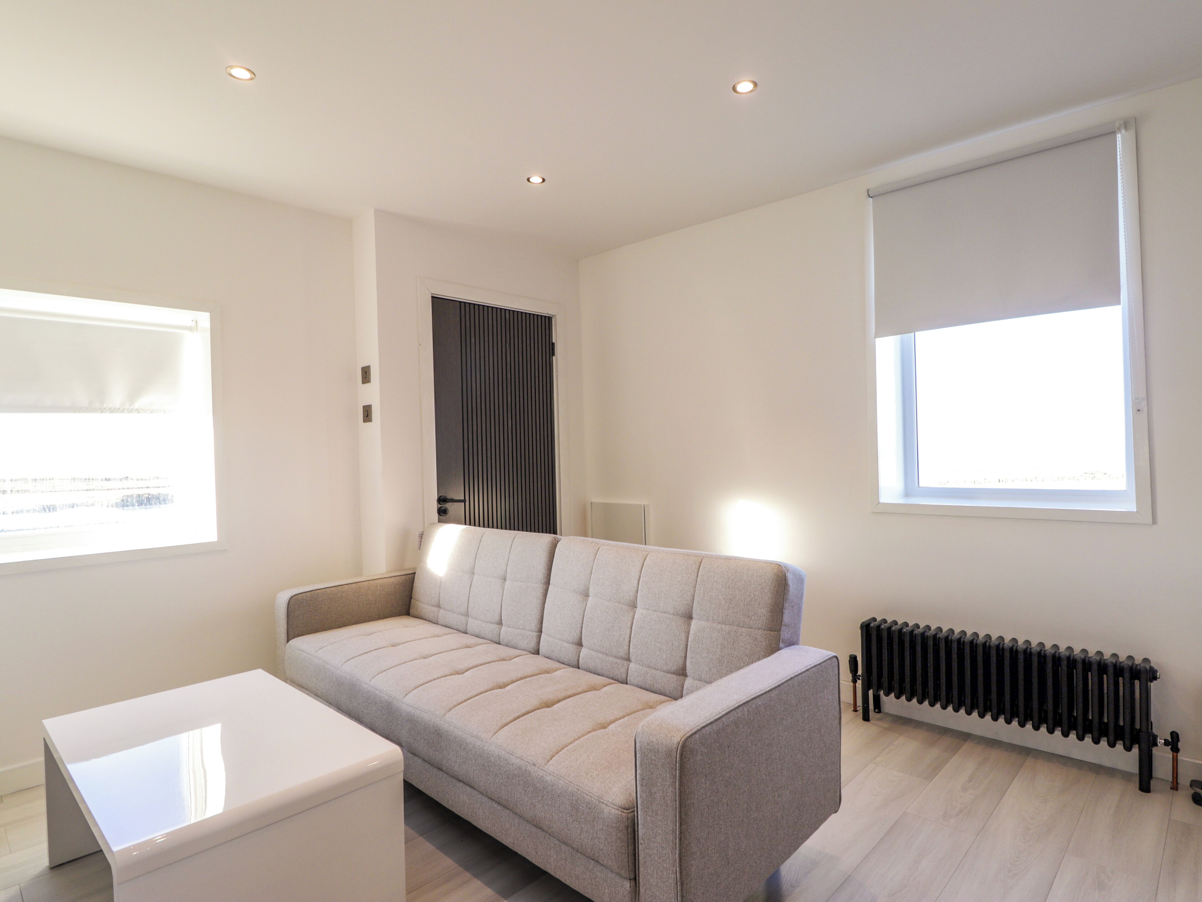 A modern living room with a sofa, a small white table, a radiator, and a window with blinds.