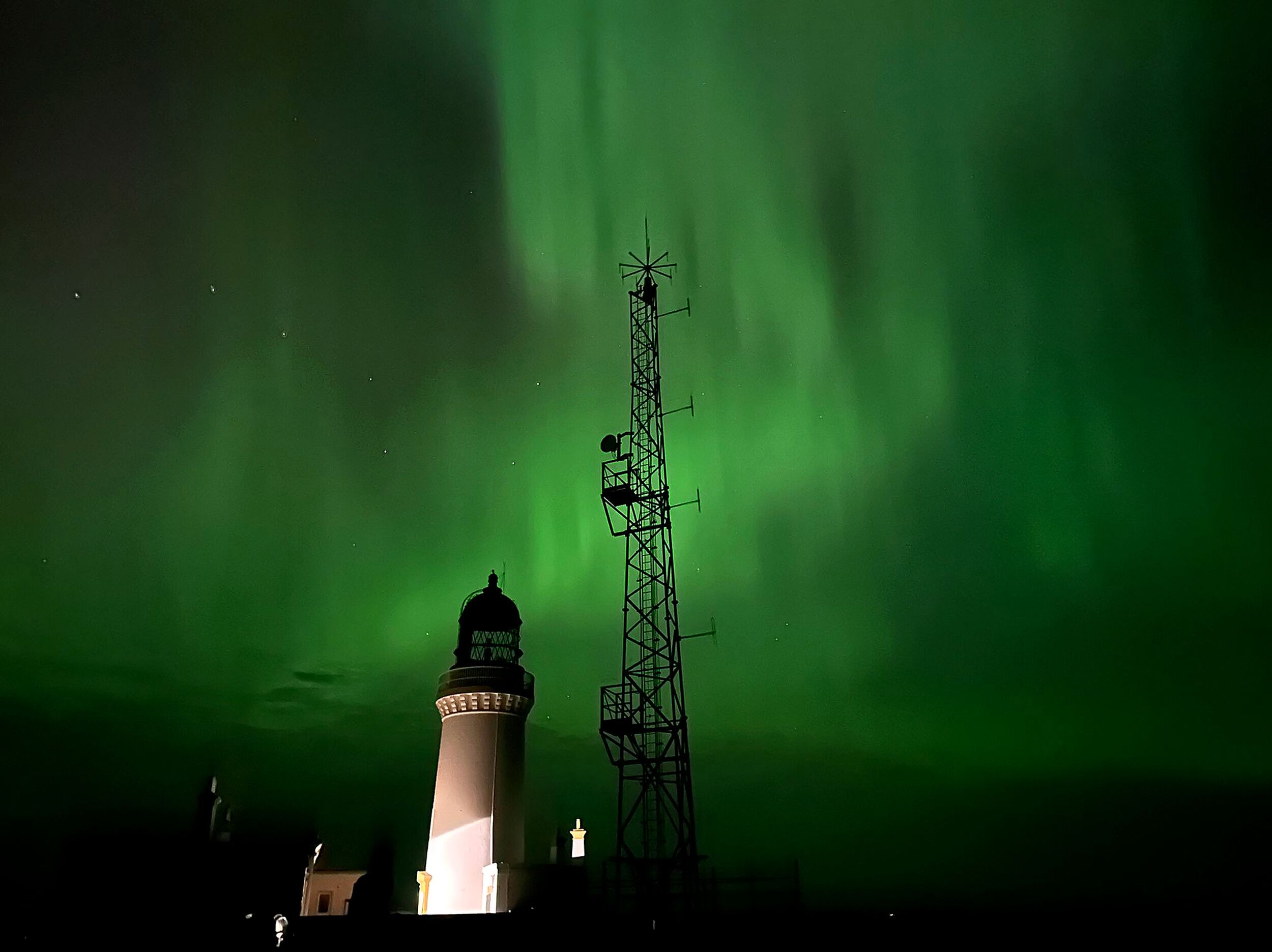A lighthouse with a white top and a dark base, a tall communication tower, and the Northern Lights in the background.