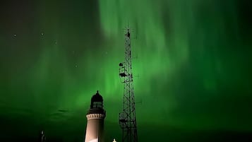 A lighthouse with a white top and a dark base, a tall communication tower, and the Northern Lights in the background.