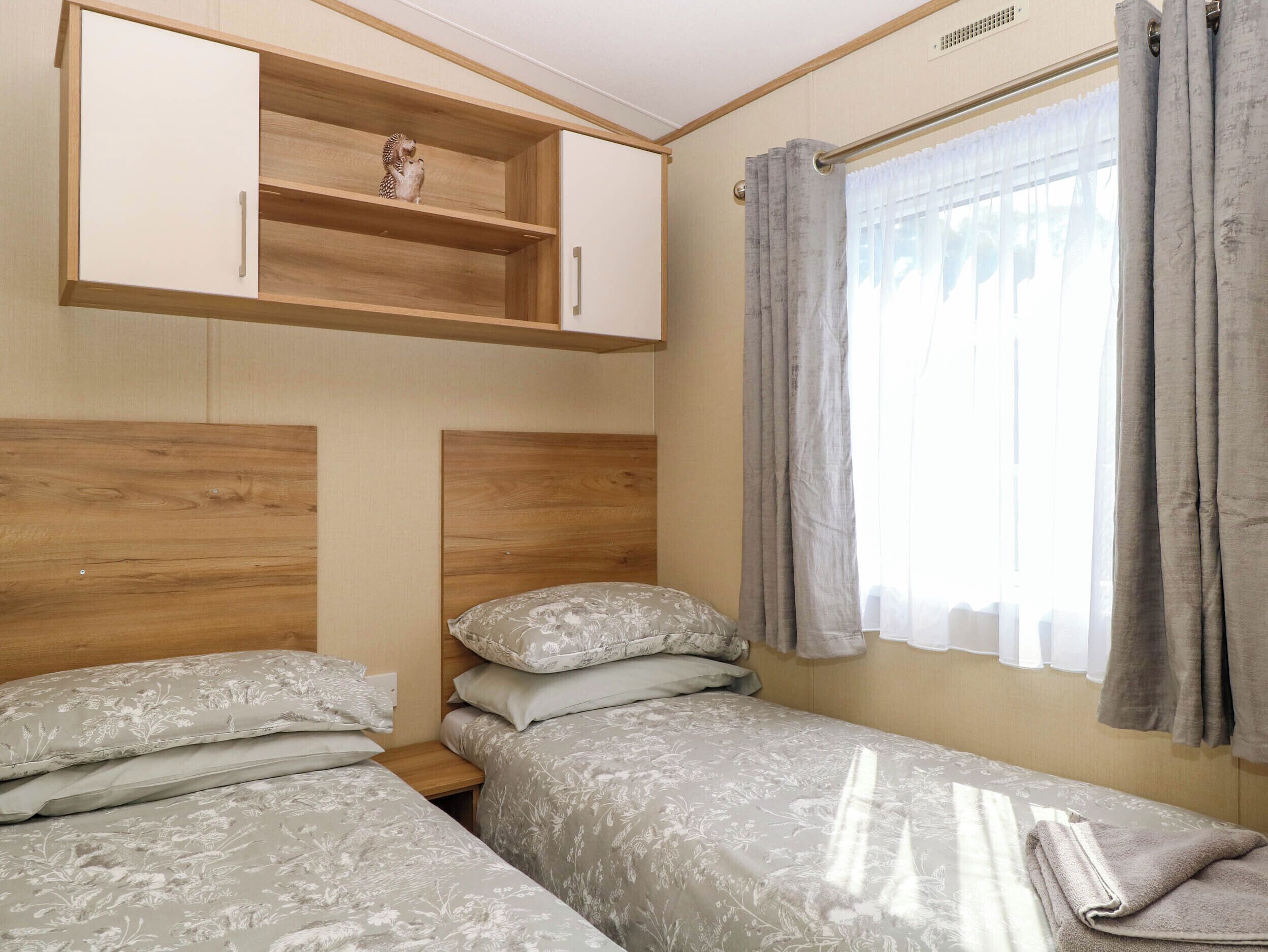A small, well-lit room with two single beds, a wooden headboard, a shelf with a small figurine, and a window with curtains.