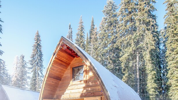 Una cabaña de madera con un jacuzzi en primer plano, rodeada de árboles cubiertos de nieve.