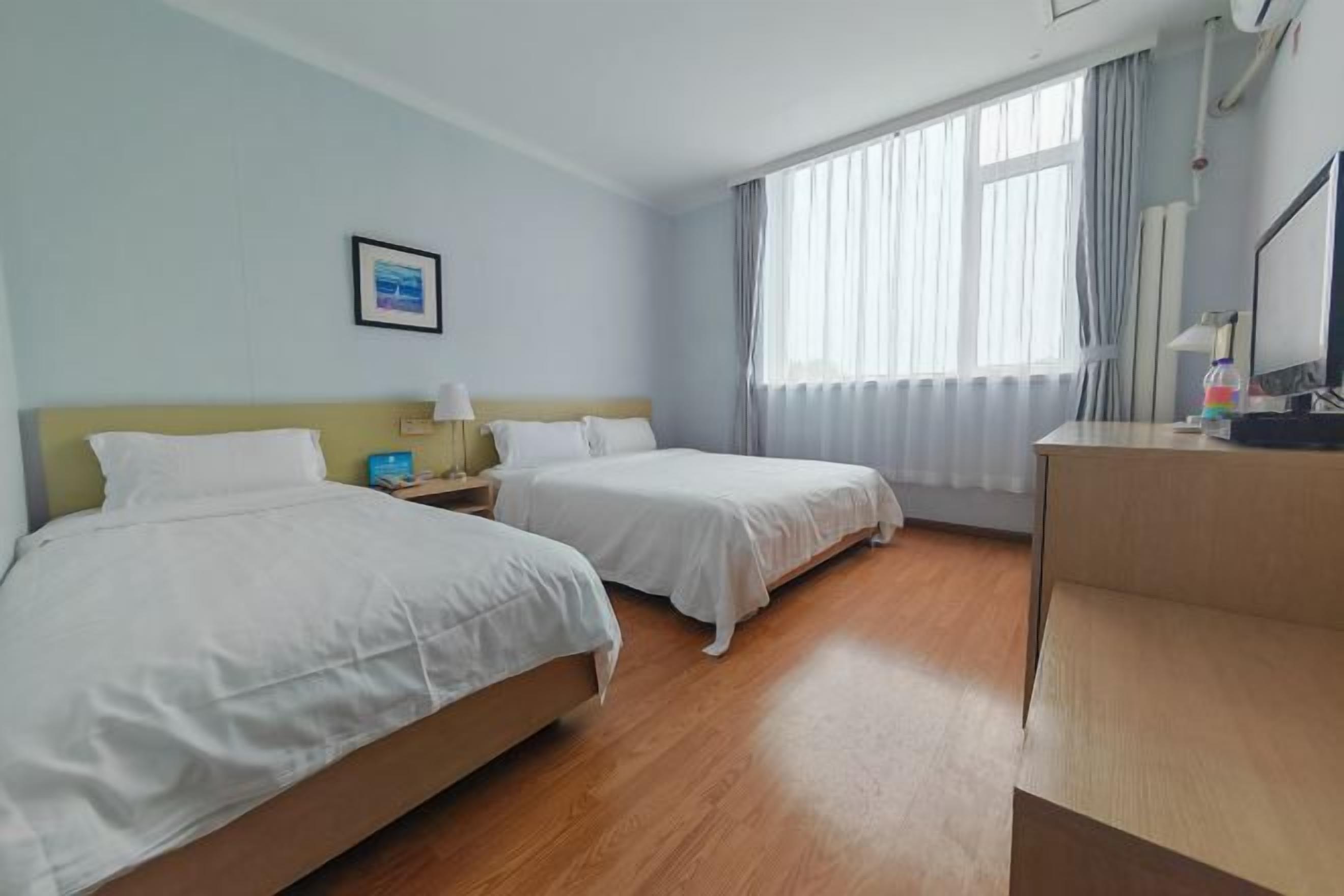 A hotel room with two beds, a desk, a chair, a nightstand, a lamp, a framed picture on the wall, and a window with curtains.