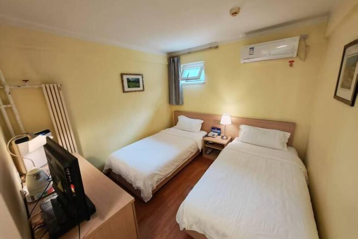 A hotel room with two single beds, a desk with a computer, an air conditioning unit, and a window with curtains.