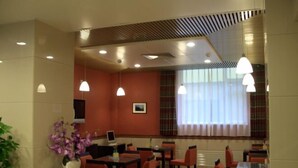 Un comedor de hotel con mesas, sillas, luces colgantes, un televisor y un ventanal con cortinas.