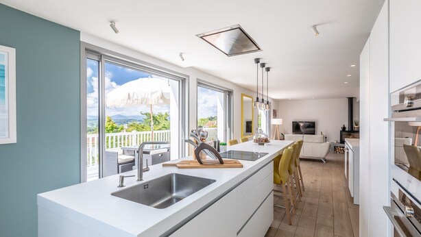 A modern kitchen with large windows, a central island, and a dining area with yellow chairs.
