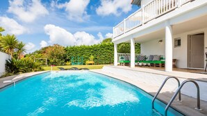 A swimming pool with a white deck, a covered seating area, and lush greenery in the background.