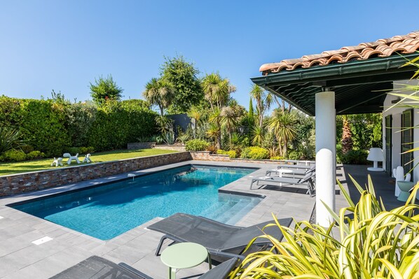 A swimming pool with lounge chairs and a small table, surrounded by greenery and a tiled roof structure.