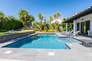 A rectangular swimming pool with a tiled deck, surrounded by palm trees and a white building with a tiled roof.