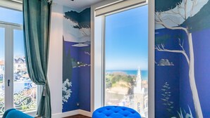 A room with a large window, a blue ottoman, and a mural on the wall. - Atalaye - Grand appartement vue mer (Biarritz)