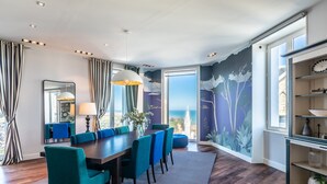 A modern dining area with a wooden table, teal chairs, and a city view through large windows. - Atalaye - Grand appartement vue mer (Biarritz)
