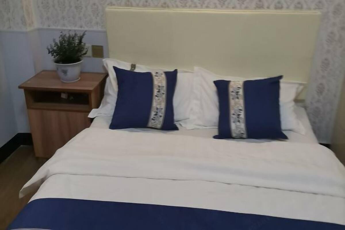 Una cama con ropa de cama blanca y azul, dos almohadas con un diseño estampado, una mesita de noche de madera con una planta en maceta y un cabecero color crema.