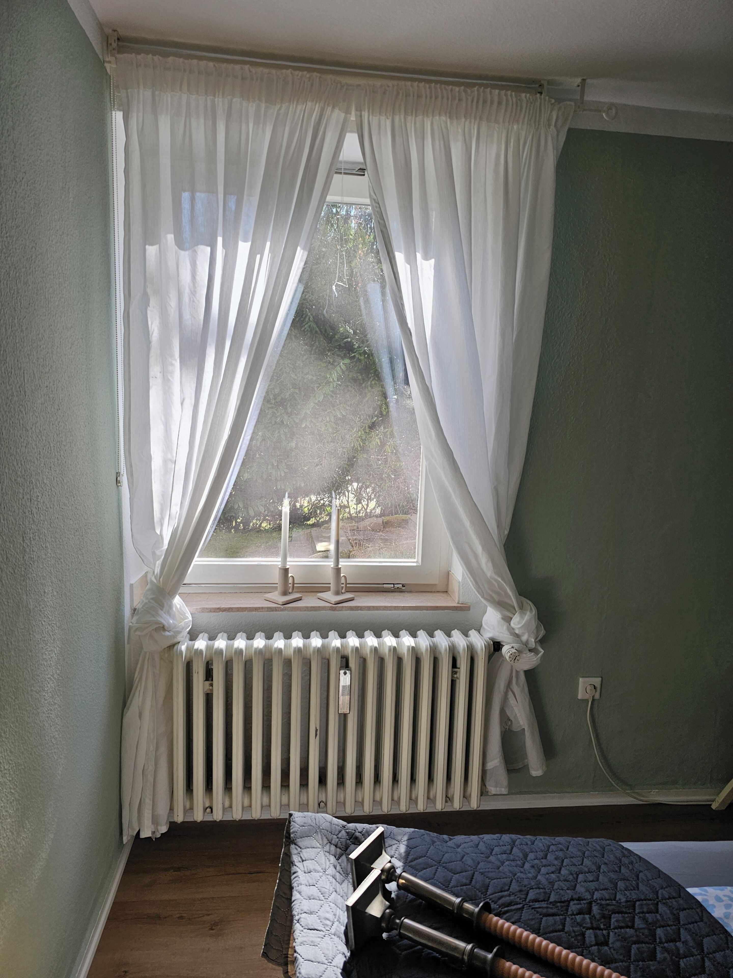 Una habitación con ventana, cortinas blancas, radiador y cama con colcha oscura.