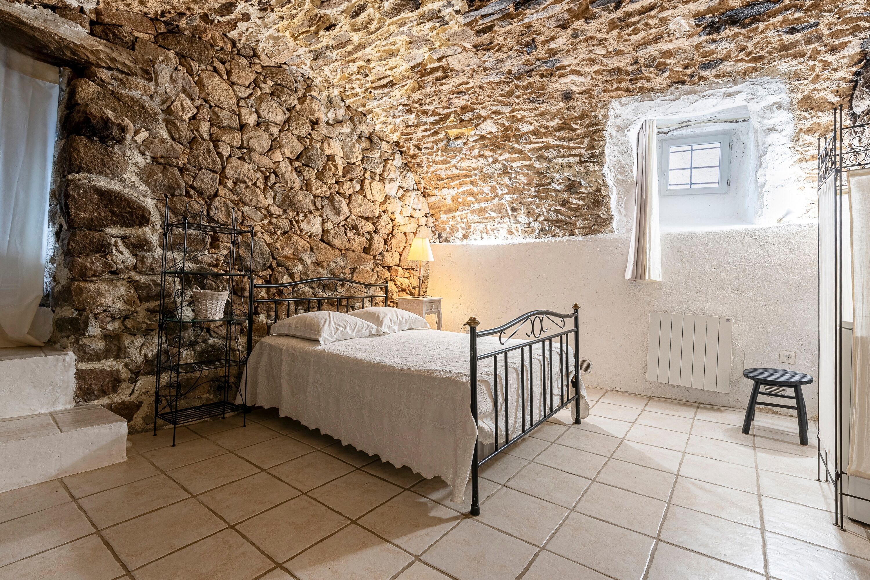 A bedroom with a stone wall, a bed, a nightstand, a window with a view, and a radiator.