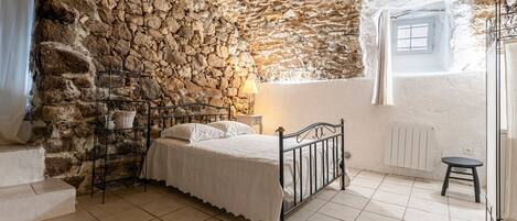 A bedroom with a stone wall, a bed, a nightstand, a window with a view, and a radiator.
