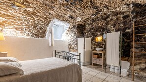 Une chambre avec un plafond en pierre, un lit recouvert d’une courtepointe blanche, une fenêtre ornée d’un rideau léger et une échelle en bois appuyée contre le mur.