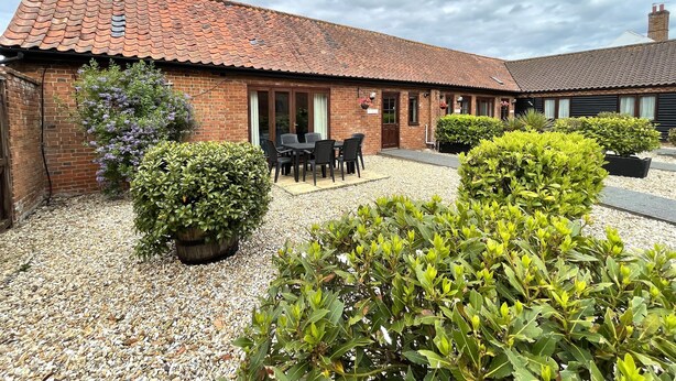 A brick building with a tiled roof, a patio with a table and chairs, and various potted plants.