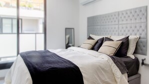 A modern bedroom with a large bed, a grey headboard, and a window with a view of buildings. - Luxury 1-bedroom apartment near WTC Montevideo with infinit pool (Montevideo)
