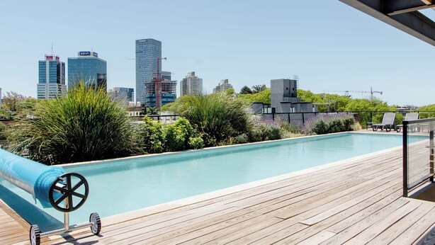 A rooftop pool with a city skyline in the background, surrounded by greenery and a wooden deck. - Luxury 1-bedroom apartment near WTC Montevideo with infinit pool (Montevideo)