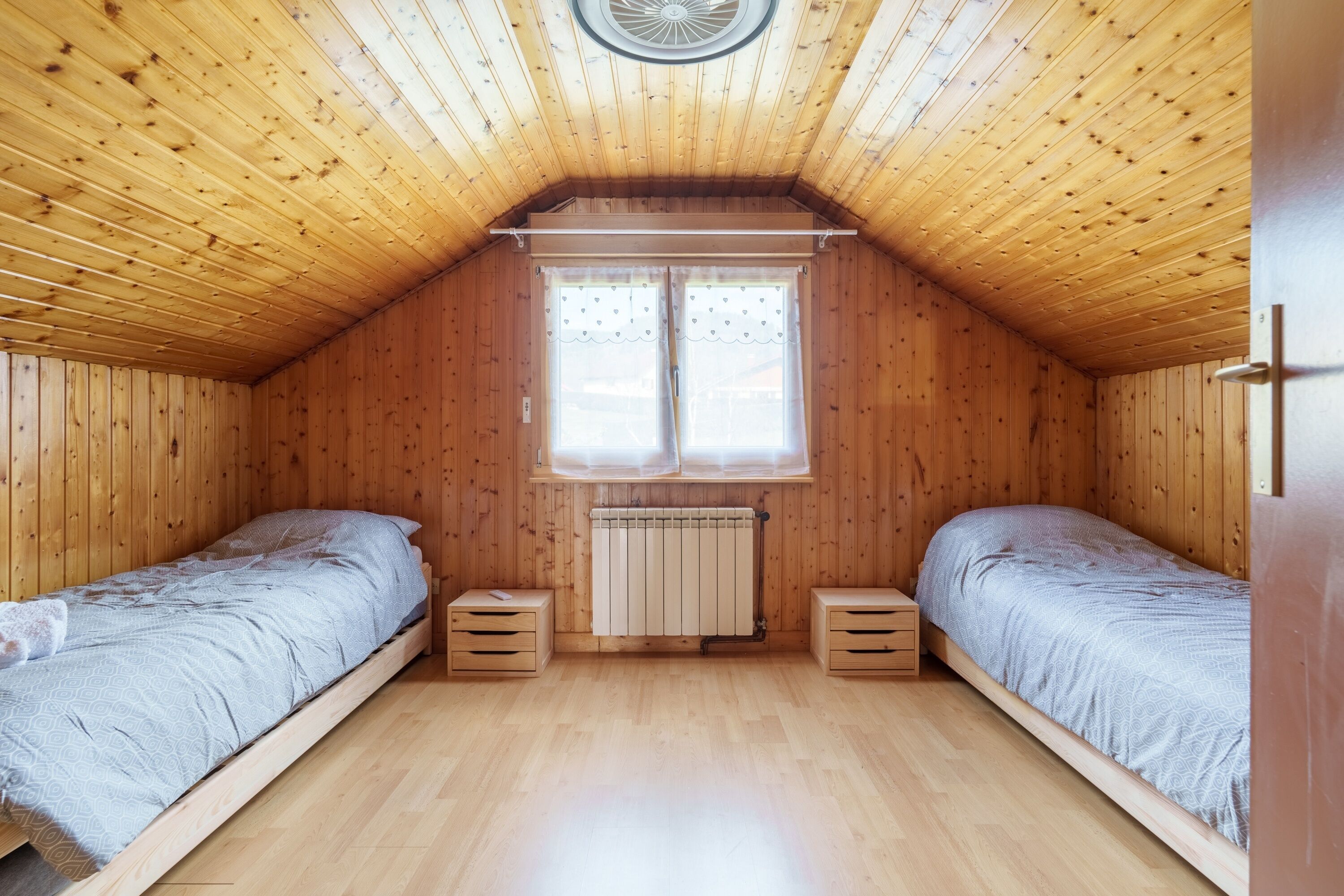 Una habitació petita amb panells de fusta, dos llits individuals, una finestra amb cortines i un radiador.