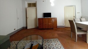 Une pièce avec un sol carrelé, un meuble en bois sur lequel est posée une télévision, une table à manger avec des chaises et un tapis à motifs.