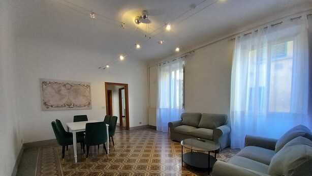 Un vaste espace de vie agrémenté d’un sol à motifs, d’un canapé, d’une table à manger avec des chaises et d’une grande fenêtre ornée de rideaux légers.