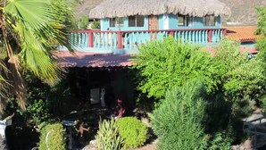Un edificio con techo de paja y fachada azul y roja, rodeado de exuberante vegetación y colinas al fondo.