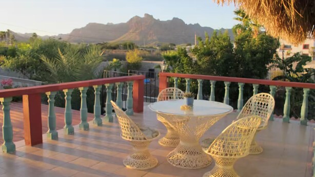 Una terraza con una mesa redonda y cuatro sillas, barandal rojo y vistas a las montañas.