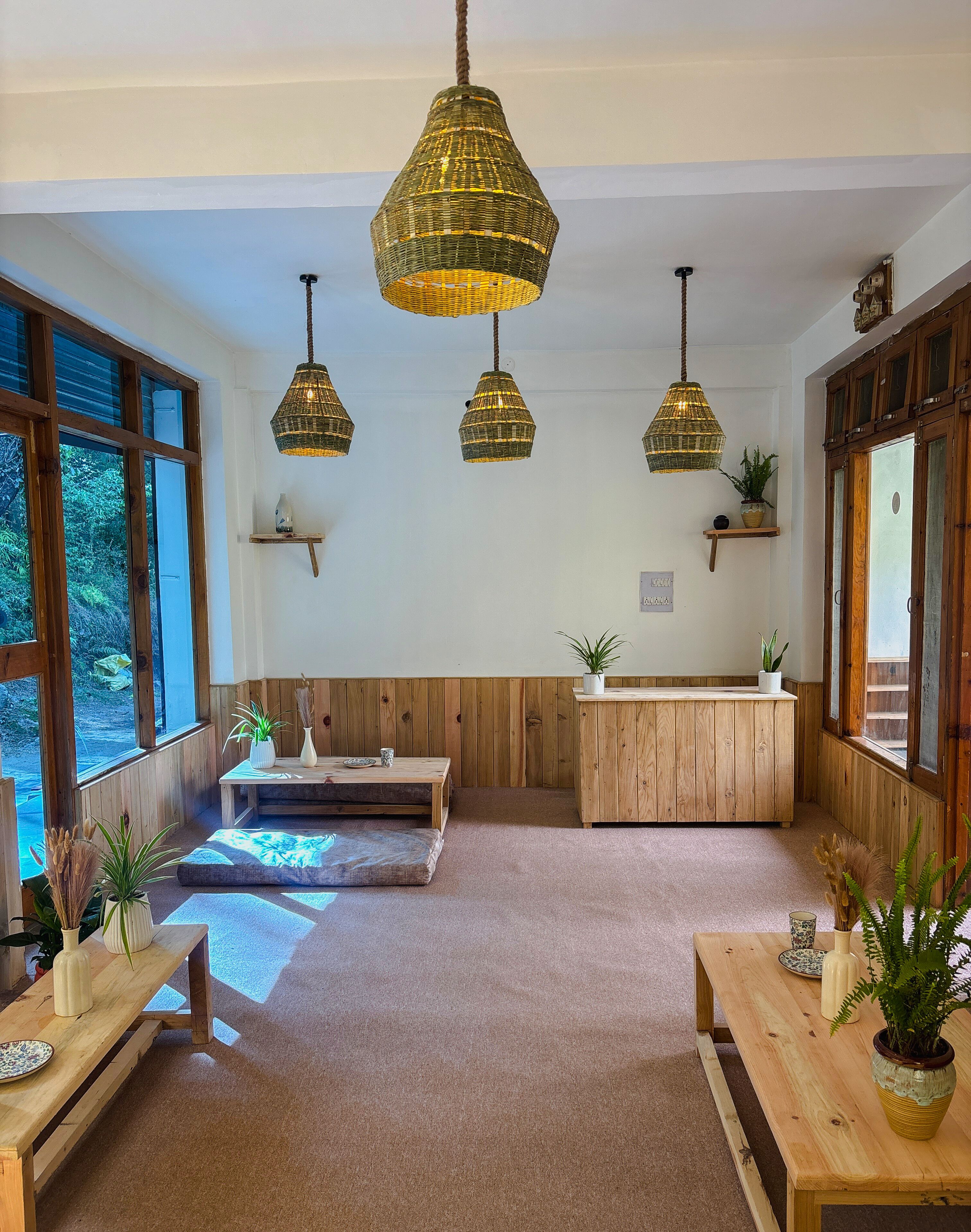 Una habitación acogedora con paredes de madera, ventanas amplias y tres lámparas colgantes de mimbre. Hay mesas y bancos de madera, y un pequeño estante con plantas.