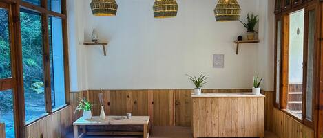 Una habitación acogedora con paredes de madera, ventanas amplias y tres lámparas colgantes de mimbre. Hay mesas y bancos de madera, y un pequeño estante con plantas.