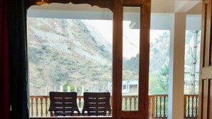 A room with a bed, a wooden floor, and a balcony with a view of a mountainous landscape. - The Woods - Tirthan Valley (Banjar)