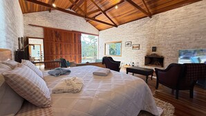 A spacious bedroom with a large bed, wooden ceiling beams, a fireplace, and a view of the outdoors.