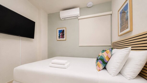 A neatly made bed with white linens and a colorful pillow, a mounted air conditioner, and a framed picture on the wall. -  Room 10: Cozy Urban Hideaway in Arts District (San Juan)