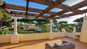 A covered patio with a wicker sofa, tiled flooring, and a view of surrounding buildings and trees.