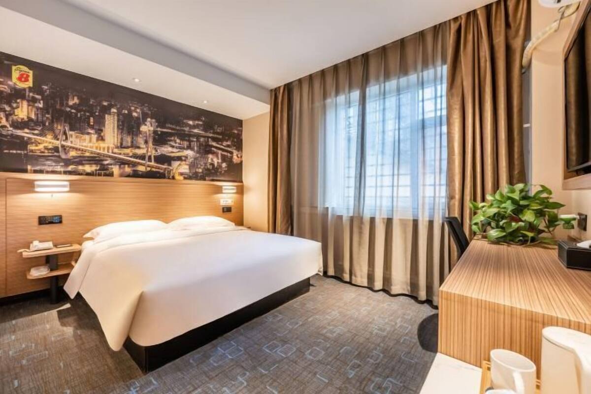 A hotel room with a large bed, a desk with a plant, and a cityscape mural on the wall.