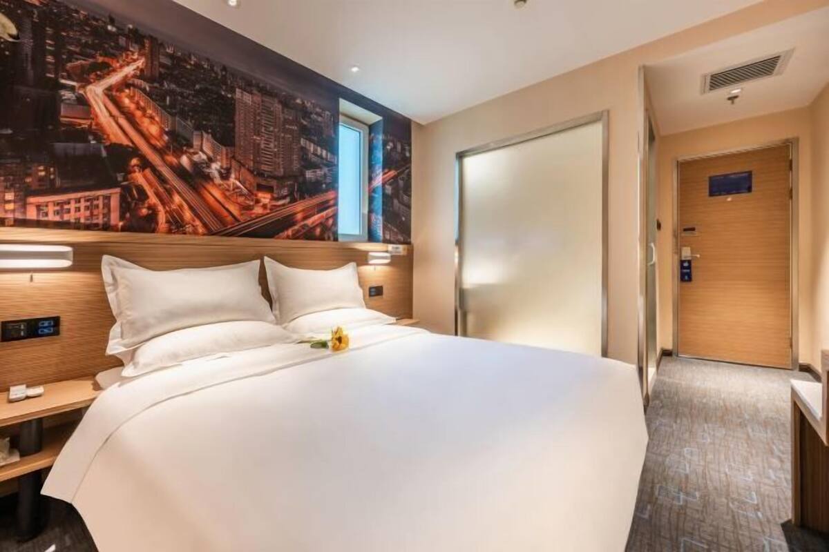 A hotel room with a large bed, a nightstand, a wooden headboard, a cityscape mural, a door, and a small table.