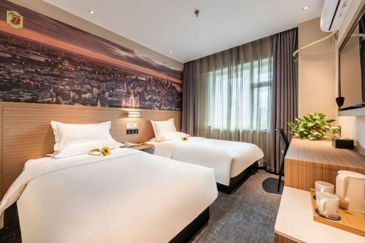 A hotel room with two beds, a desk with a chair, a large window with curtains, and a wall mural.