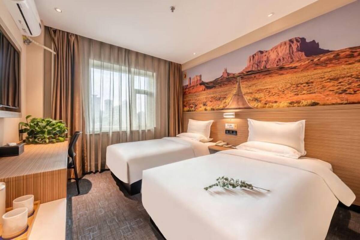 A hotel room with two beds, a desk with a chair, a plant, and a desert-themed mural on the wall.