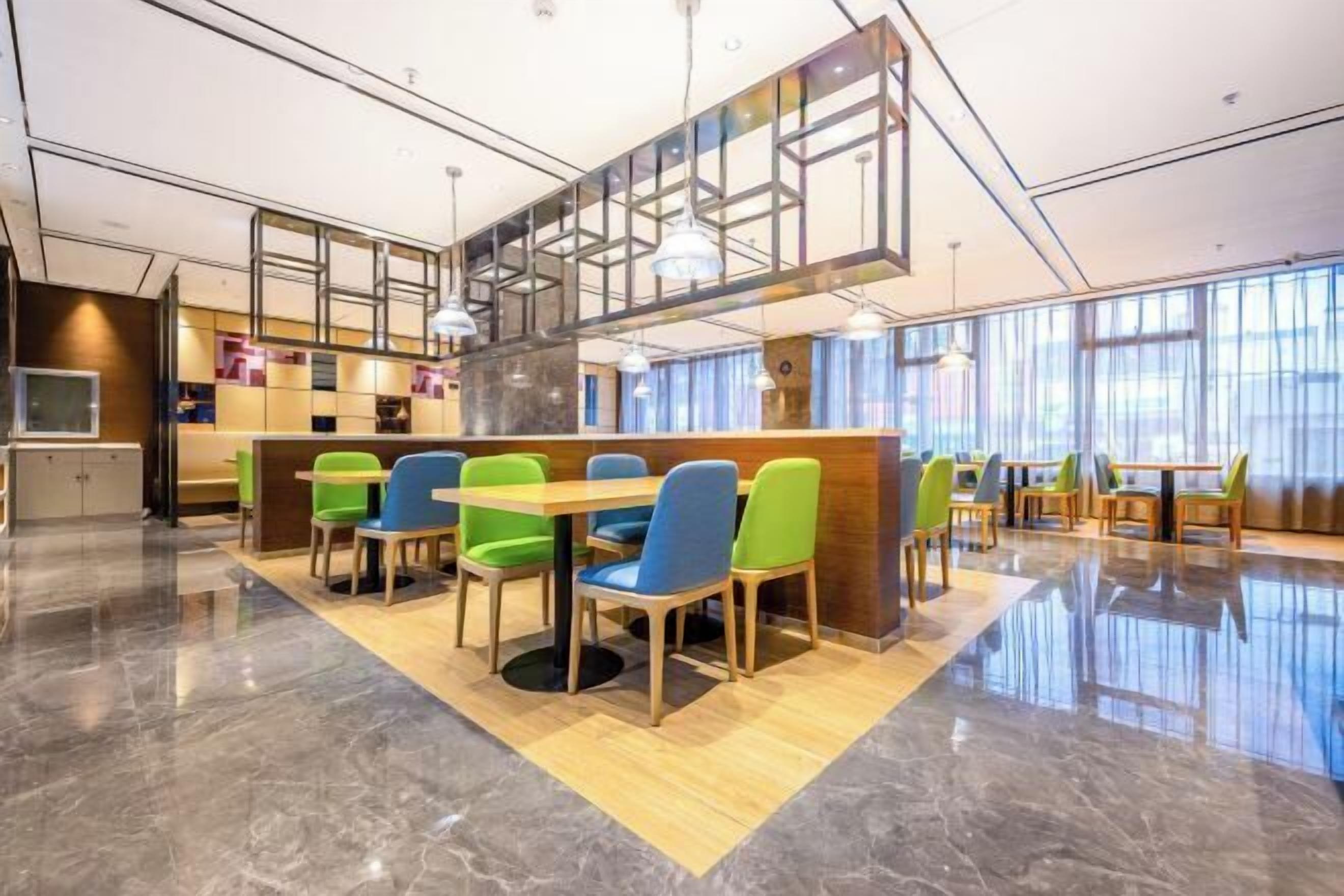 A modern dining area with a central wooden table surrounded by colorful chairs, a high ceiling with exposed beams, and large windows providing natural light.