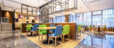 A modern dining area with a central wooden table surrounded by colorful chairs, a high ceiling with exposed beams, and large windows providing natural light.