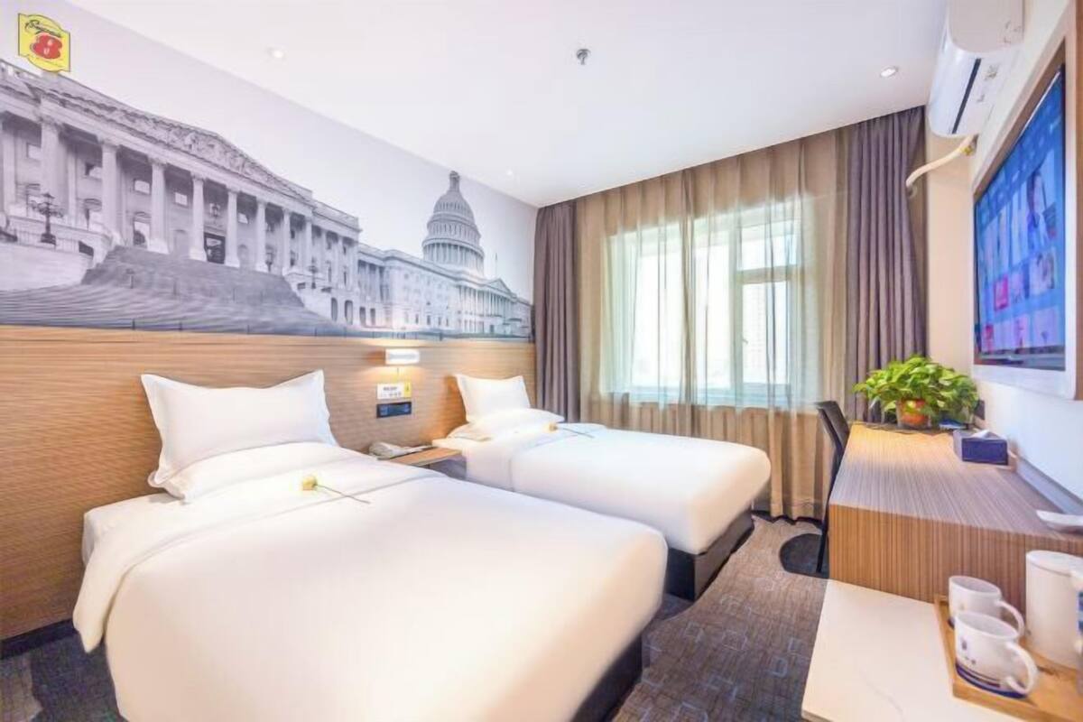 A hotel room with two beds, a desk, a chair, a potted plant, and a large mural of a building.