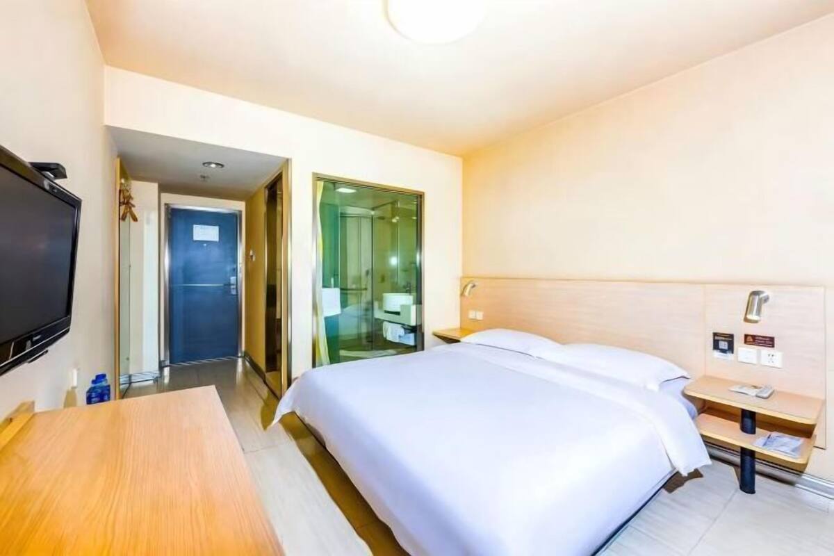 A hotel room with a bed, a TV, a desk, and a bathroom visible through a glass door.