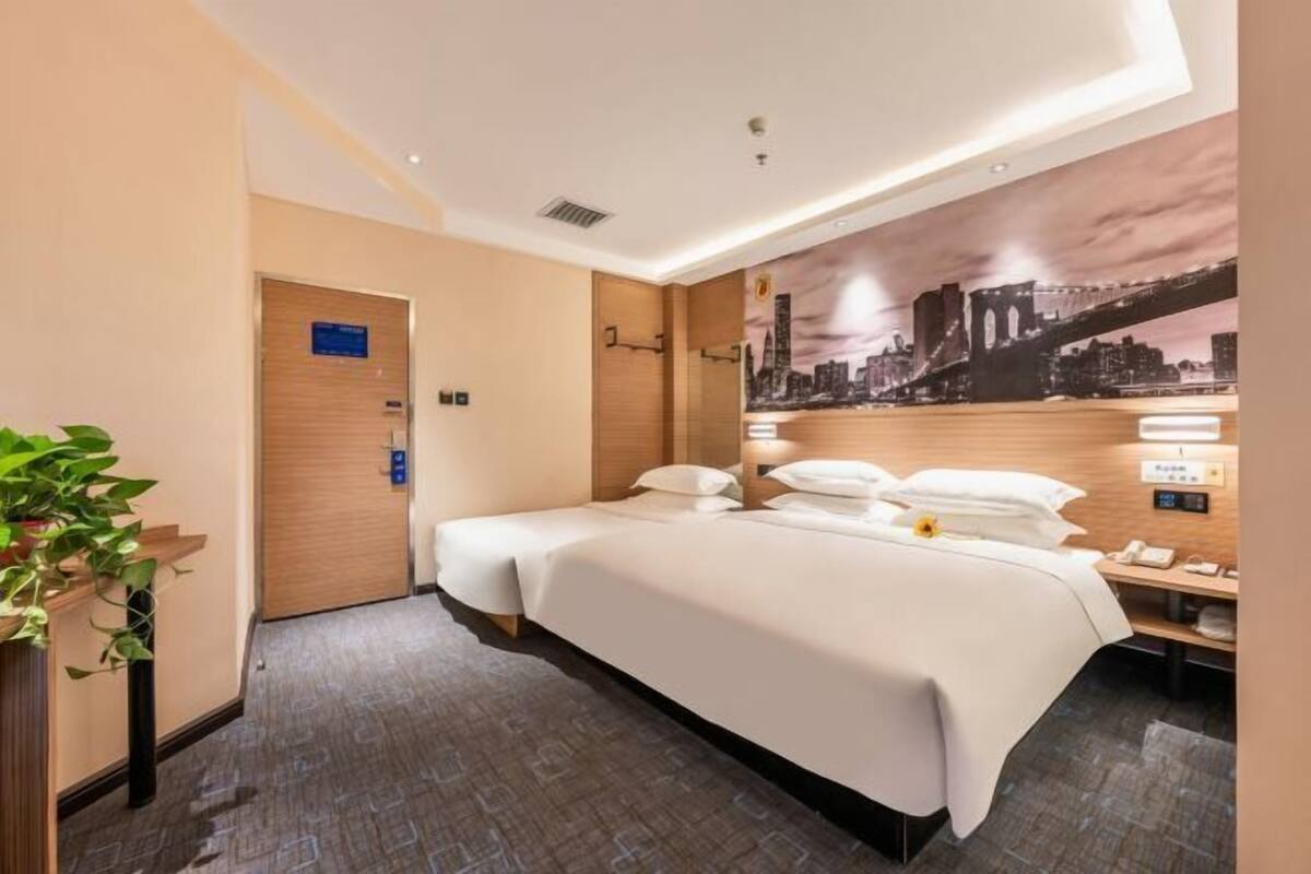 A hotel room with a large bed, a cityscape mural on the wall, a plant, and a wooden door.